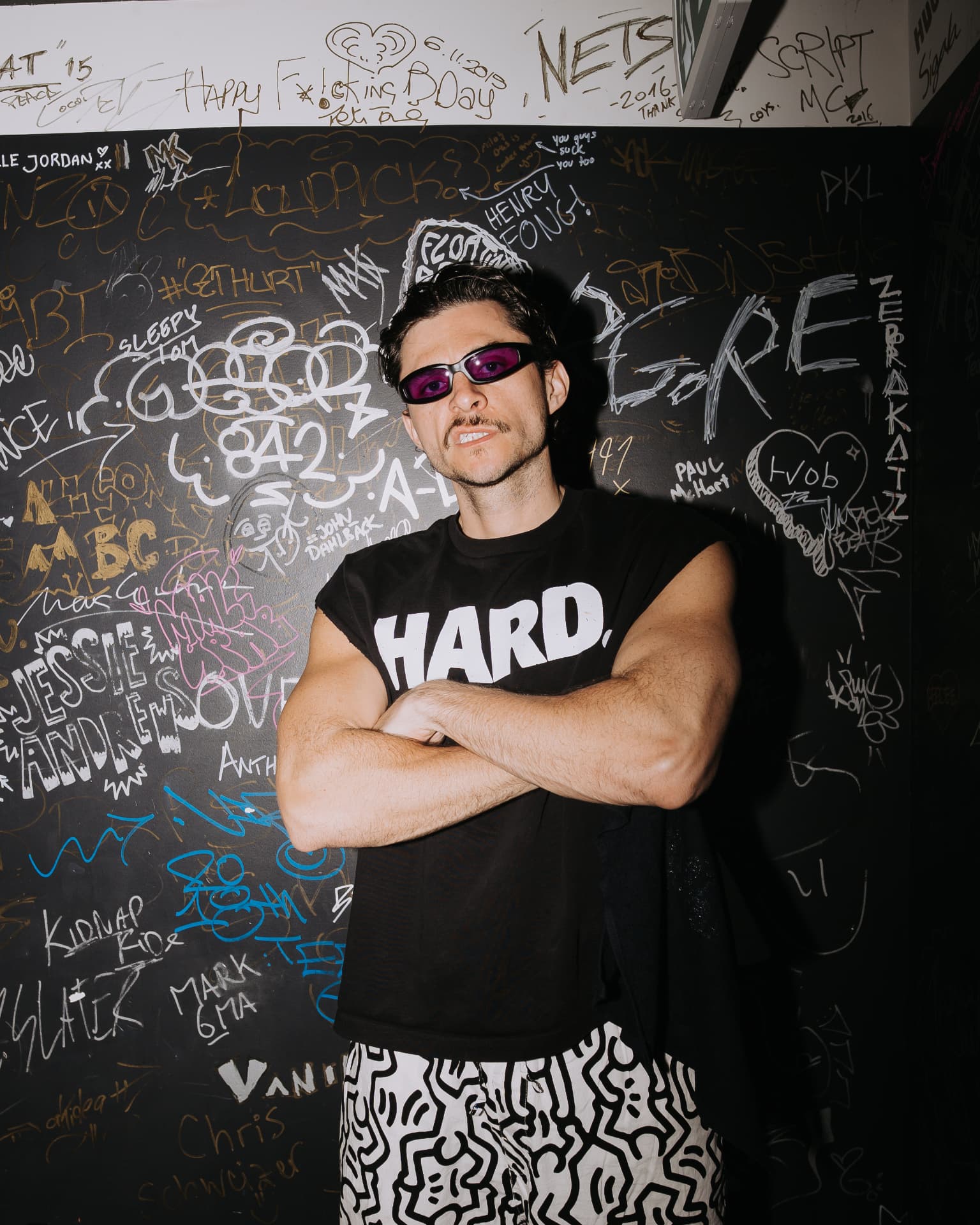 Jake Shore in purple sunglasses with arms crossed in front of a signature-covered wall
