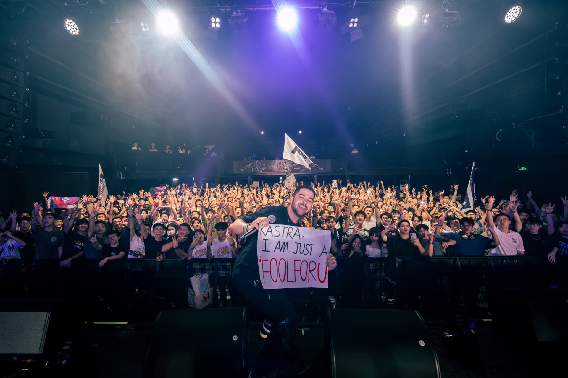 Kastra on stage facing a packed crowd holding signs at a live show